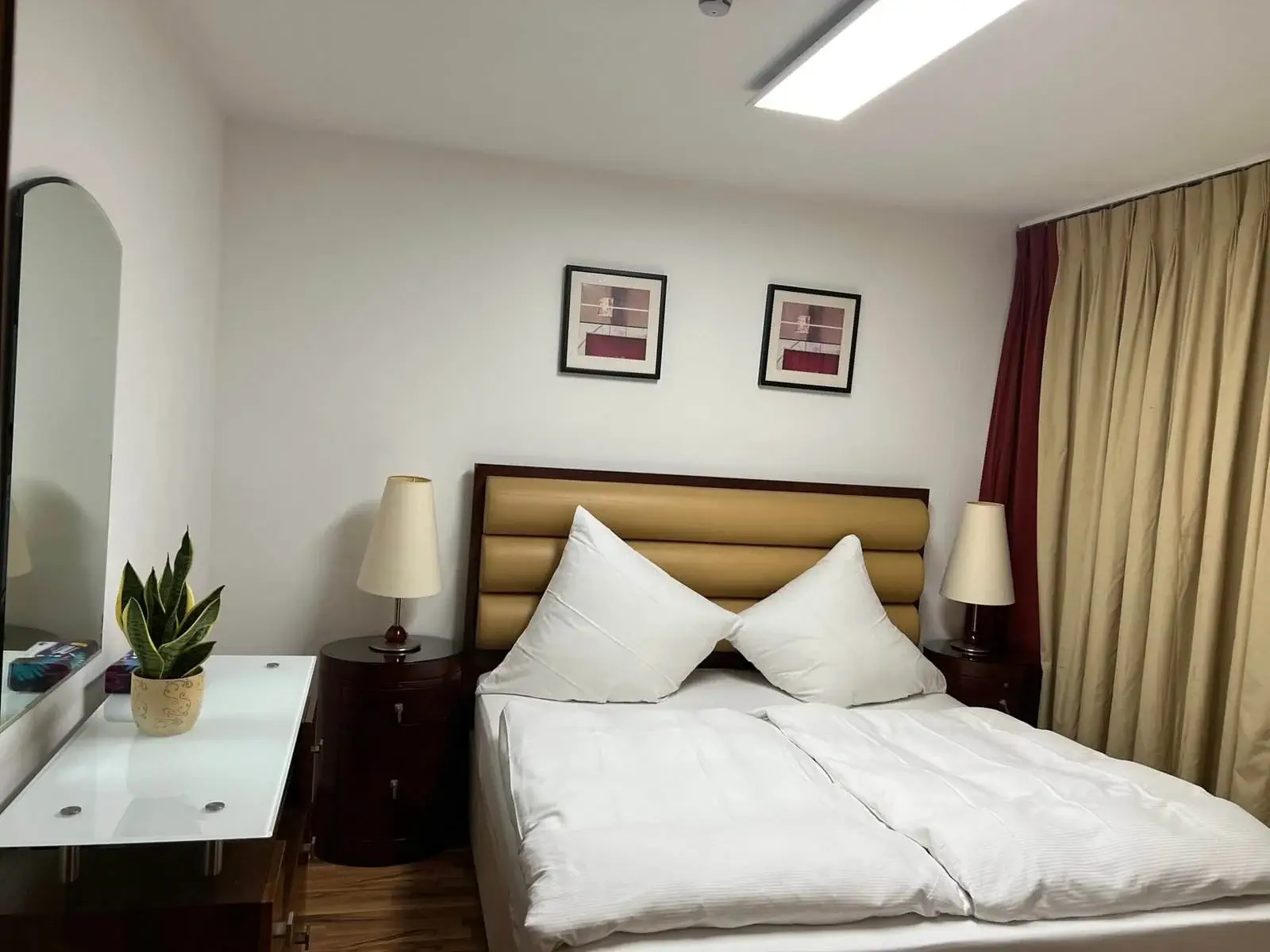 Ein gemütliches Schlafzimmer mit einem Doppelbett mit weißer Bettwäsche und großen Kissen. Zwei hölzerne Nachttische mit Lampen flankieren das Bett. Über dem Bett hängen zwei abstrakte Gemälde. Links steht eine Kommode mit einer Topfpflanze und einem großen Spiegel. Beige Vorhänge verdecken ein Fenster rechts.