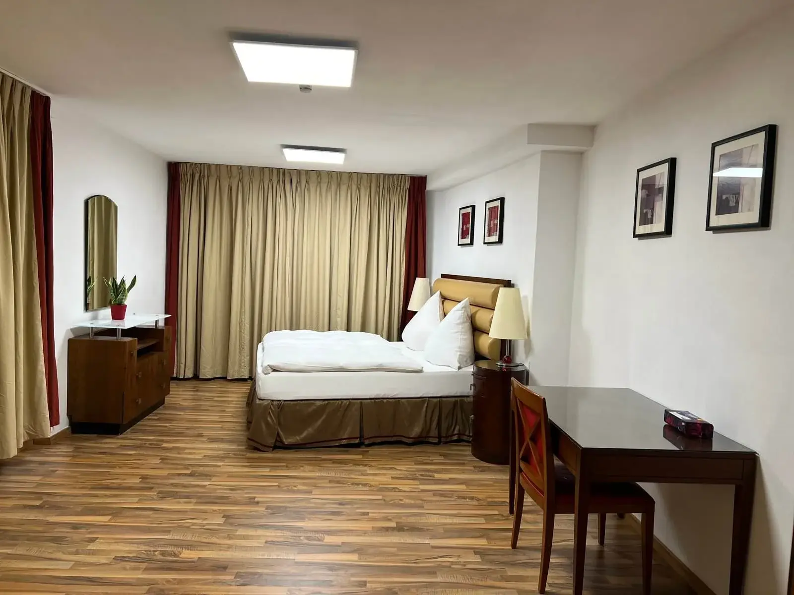 Geräumiges Hotelzimmer mit Holzboden, einem Doppelbett mit weißer Bettwäsche und gepolstertem Kopfteil. Rechts steht ein Schreibtisch mit Stuhl, links eine Kommode mit Topfpflanze und Spiegel. Cremefarbene Vorhänge verdecken das Fenster, und gerahmte Kunstwerke schmücken die weißen Wände.